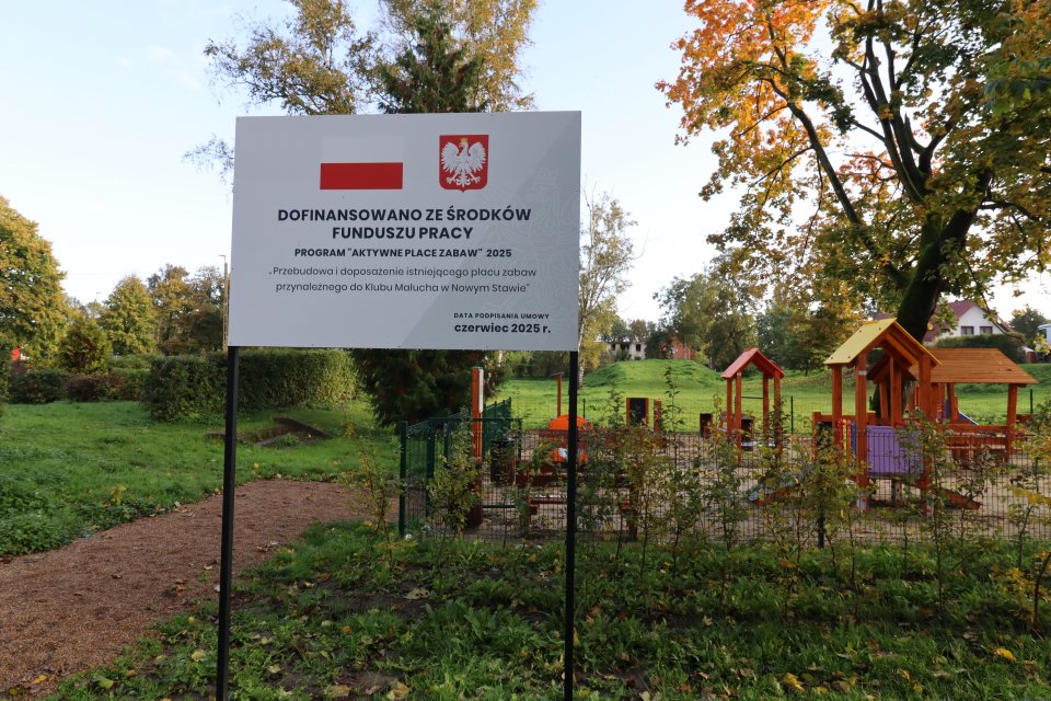 Tablica informująca o dofinansowaniu - biała tablica a na niej od góry biało-czerwona flaga i godło Polski. Poniżej informacja skąd pochodzi dofinansowanie oraz nazwa zadania. W prawym dolnym rogu data podpisania umowy. W tle plac zabaw