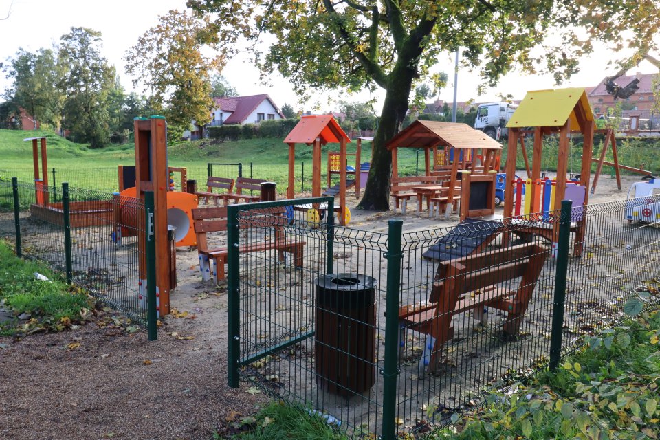 Ogrodzony plac zabaw. Widać ławki, wiaty, i elementy zabawowo-rekreacyjne,